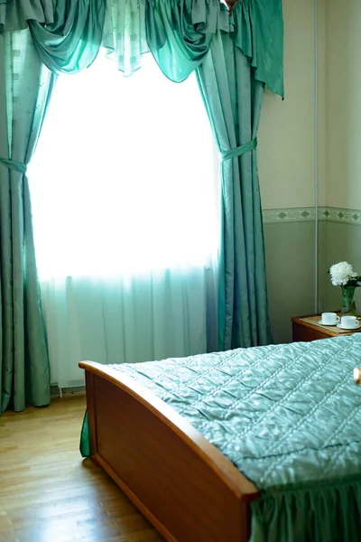 window with green curtains in the bedroom with a bed, bedside table and cups