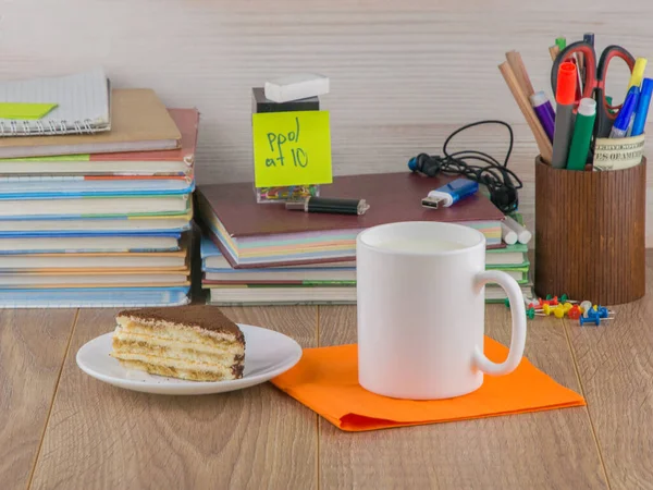 high schoolboy: a mug of milk on an orange napkin and a piece of cake on a plate, on the desktop, front view, close