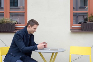 Adam kafenin yakınındaki bir masada oturuyor, cep telefonunda bir şey arıyor, masanın üstünde yalan makineleri.
