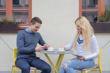 Genç bir adam ve uzun saçlı sarışın bir kız kahve ya da çay içerler masadaki bir kafenin yanında, bir adam bir kıza telefonundan bir şey gösterir, gülerler.