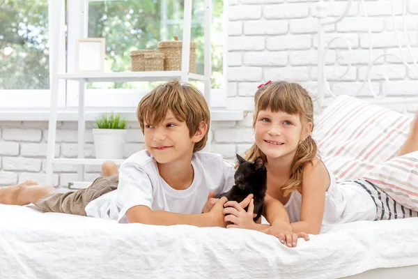 Zwei kleine glückliche Kinder spielen mit schwarzer Katze auf weißem Bett bei hom — Stockfoto