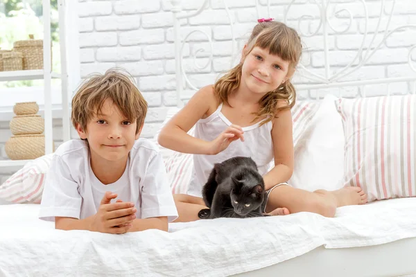Duas crianças felizes brincando com gato preto na cama branca no hom — Fotografia de Stock