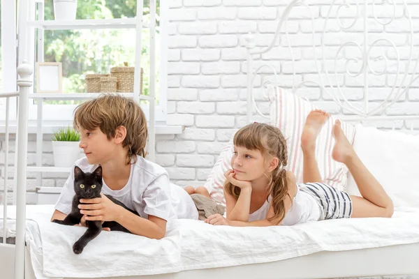 Drei kleine glückliche Kinder spielen mit schwarzer Katze auf weißem Bett um h — Stockfoto