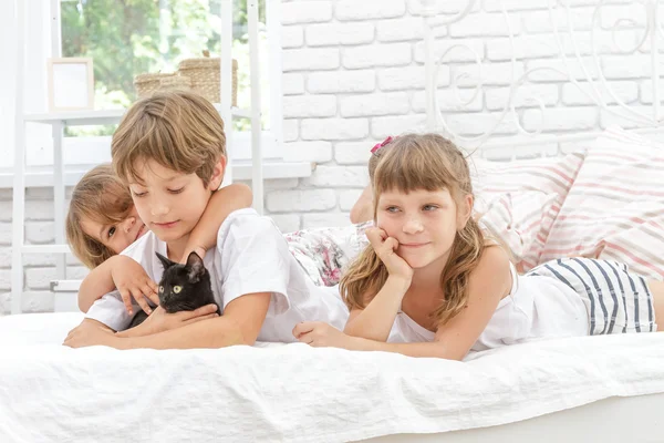 Tři malé šťastné děti hrají s černou kočkou na bílé posteli v h — Stock fotografie