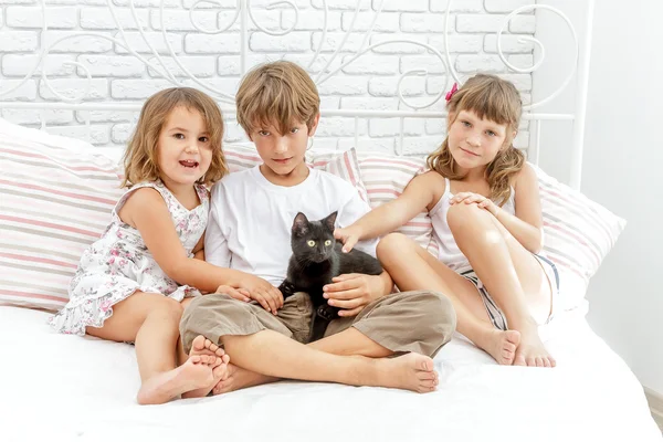 Tres pequeños niños felices jugando con gato negro en la cama blanca en h —  Fotos de Stock