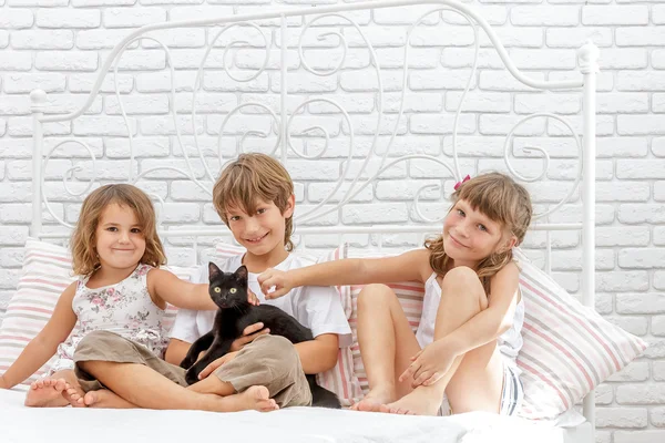 Três crianças pequenas felizes que jogam com gato preto na cama branca em h — Fotografia de Stock
