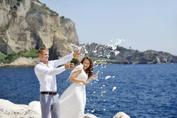 Young married couple uncorking champagne bottle by the sea after their wedding — Stock Photo, Image