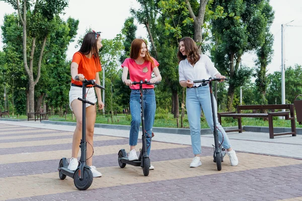 E-scooter, electric scooter, Ecological urban city transport, alternative energy, alternative lifestyle. Three Young girl friends on vacation having fun driving electric scooter through city park