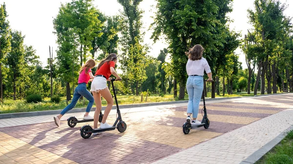 E-scooter, electric scooter, Ecological urban city transport, alternative energy, alternative lifestyle. Three Young girl friends on vacation having fun driving electric scooter through city park