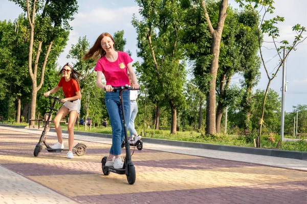 E-scooter, electric scooter, Ecological urban city transport, alternative energy, alternative lifestyle. Three Young girl friends on vacation having fun driving electric scooter through city park