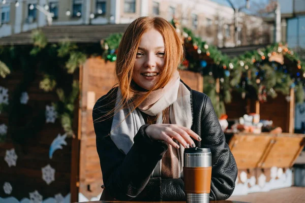 Молодая рыжая женщина, наслаждающаяся солнцем и пьющая кофе, прогуливается по улицам зимнего города. Счастливая молодая улыбающаяся женщина пьет горячий напиток на рождественской ярмарке, рынке — стоковое фото