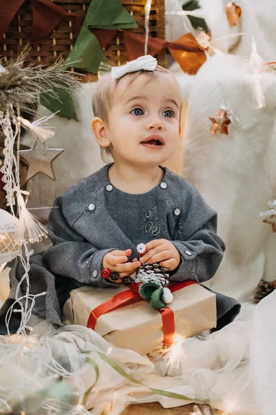 Симпатичная маленькая улыбчивая девочка с коробкой подарков у новогодней елки дома. Маленькая улыбающаяся девочка на фоне рождественской гирлянды и подарочных коробок с ленточкой — стоковое фото