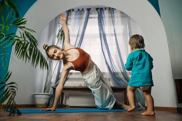 Home Fitness, Training im neuen Normalzustand, Pandemiezeit, Yoga zu Hause. Mutter trainiert, praktiziert Yoga mit Baby-Mädchen im selben Raum — Stockfoto