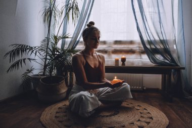 Yerde oturan genç bir kadın, mumlar yakıyor, meditasyonun tadını çıkarıyor, evde yoga egzersizi yapıyor. Zihinsel sağlık, kişisel bakım, stres yok, sağlıklı alışkanlık, farkındalık yaşam tarzı, endişe giderme konsepti