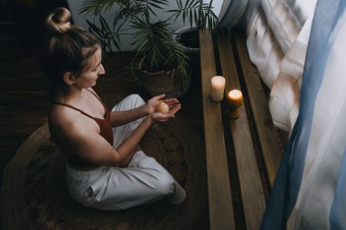 Yerde oturan genç bir kadın, mumlar yakıyor, meditasyonun tadını çıkarıyor, evde yoga egzersizi yapıyor. Zihinsel sağlık, kişisel bakım, stres yok, sağlıklı alışkanlık, farkındalık yaşam tarzı, endişe giderme konsepti