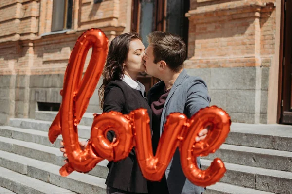 San Valentín celebración del día y citas concepto. Feliz pareja amorosa con globos rojos de amor en la calle de la ciudad. Retrato exterior de pareja joven con globo de palabra Amor. —  Fotos de Stock