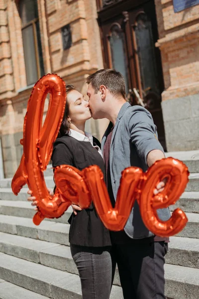 San Valentín celebración del día y citas concepto. Feliz pareja amorosa con globos rojos de amor en la calle de la ciudad. Retrato exterior de pareja joven con globo de palabra Amor. —  Fotos de Stock