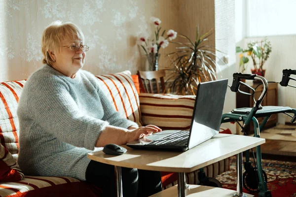 Senior mature candid real Caucasian woman look at laptop screen at home. Happy senior blonde female have online video call with family via internet. Elderly technology concept.