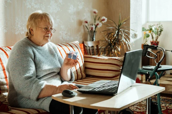 Senior mature candid real Caucasian woman look at laptop screen at home and hold gift box. Happy senior blonde female have online video call with family via internet. Elderly technology concept.