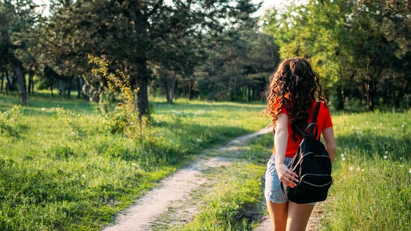 Local travel, Rural tourism, Staycations, gen z traveler, Young Adventurer, Solo Travel. Back view of Young brunette woman with backpack walking on country road to forest