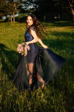 Parti mezuniyet balosu konsepti. Mavi balo elbiseli, esmer ve güzel bir kadın. Arka planda leylak demeti var. Uzun gece elbisesi içindeki esmer kızın açık hava romantik portresi.