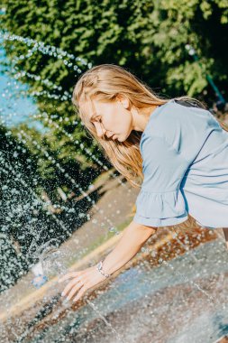 Sıcak yaz, sıcakta serinlemek. Şehir parkındaki fıskiyede mavi elbiseli genç bir kadın. Sıcak bir günde ne giyilir?