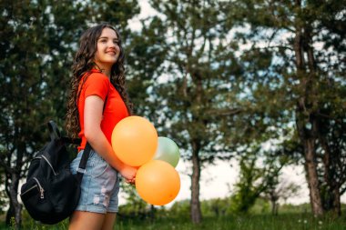 Yaz tatili, yaz tatilinde okul tatili, öğrenciler tatil, akademik yıl sonu. Sırt çantası ve renkli balonları olan genç esmer kız yazın tadını çıkarıyor.