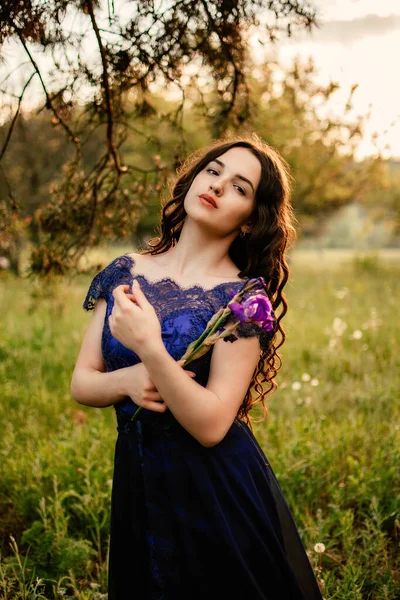 Party graduation prom fairytale concept. Beautiful brunette young woman in blue prom dress with iris flower on nature background. Outdoor romantic portrait of brunette girl in long evening dress