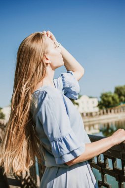 Yazı şehirde geçirmek, sıcağa dayanmak, yaz kıyafetleri, solunabilir kumaşlar. Şehir caddesinde mavi keten elbiseli genç sarışın kadın.