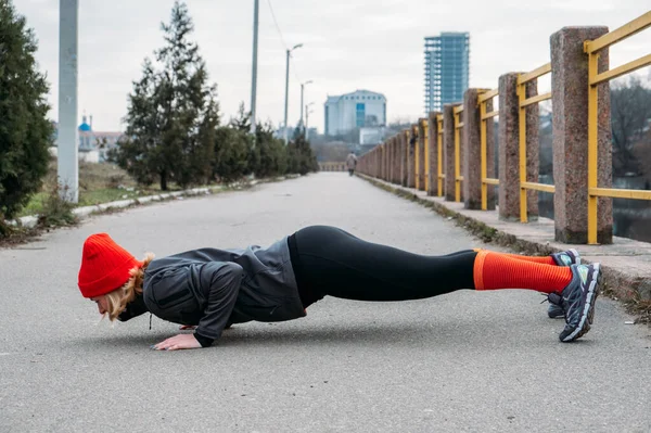 Dopasowany sportowy blondynka w średnim wieku 40 kobieta w czerwonej czapce Stojąc w Plank, robi trening, rozciąganie, korzystając fitness na ulicy miasta wiosną jesienią dzień. — Zdjęcie stockowe