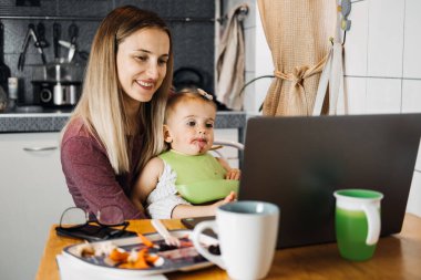 Working mother work remotely. Remote Work from home. Young mother with toddler baby girl working at home using laptop on kitchen background