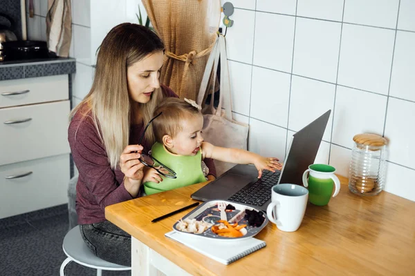 Работающая мать работает удаленно. Удаленная работа из дома. Молодая мать с маленькой девочкой работает дома, используя ноутбук на кухонном фоне — стоковое фото