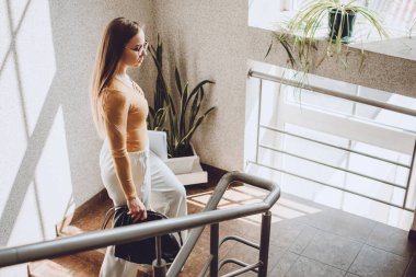 Young professionals, traineeship, practical experience. Young business woman walking up the stair in office. Young beautiful woman with handbag and documents walking indoors