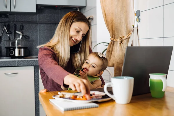 Working mother work remotely. Remote Work from home. Young mother with toddler baby girl working at home using laptop on kitchen background — Stock Photo, Image