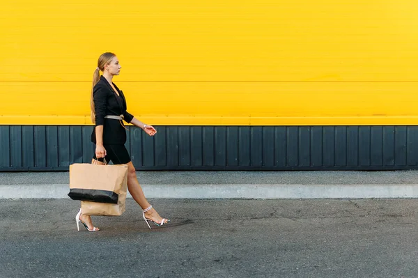 Αγορές, πωλήσεις. Κομψό μοντέρνα νεαρή γυναίκα με τα πόδια και κρατώντας τσάντες ψώνια σε κίτρινο φόντο τοίχο εξωτερική. — Φωτογραφία Αρχείου