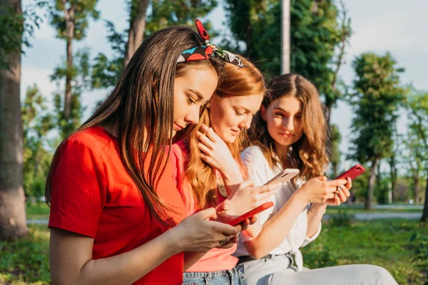 Three girl friends watching mobile smart phones and having fun sitting on bench in public park. Gen z, Teenagers devices addiction, Digital detox, youth culture, tech, social friendship