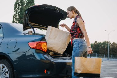 Alışveriş, indirim alışveriş yaptıktan sonra alışveriş torbalarıyla araba açan mutlu, şık, sarışın kadın. Kız alışveriş çantalarını arabaya koyuyor.
