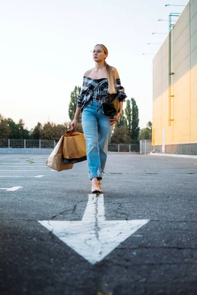 Shopping, vendita, Happy giovane donna bionda elegante con borse della spesa andare in auto nel parcheggio sulla freccia bianca dopo lo shopping. Donna con shopping bag sul parcheggio esterno. — Foto Stock