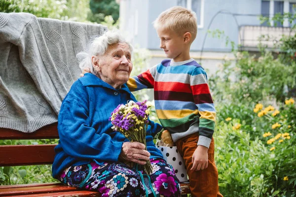 Dia dos avós, família reunida, união. A avó idosa abraça o neto ao ar livre. Neto faz surpresa para a avó e deu-lhe flores — Fotografia de Stock