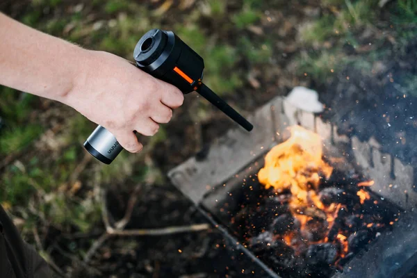 Metal bir yangını körüklemek için pille çalışan bir üfleyici kullanan bir elin yakın çekimi. Yangın başlatma teknolojisi, taşınabilir üfleyiciler, kamp aletleri, açık havada yenilik. Yüksek kalite fotoğraf