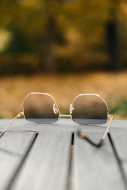 Bir çift altın çerçeveli güneş gözlüğü bulanık bir sonbahar arka planına sahip gri ahşap bir masada dinleniyor. Sonbahar sembolizmi, mevsimsel geçişler, görsel metafor, şiirsel hikaye anlatımı. Yüksek kalite fotoğraf