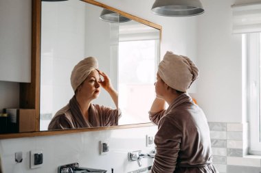 A woman in a robe and hair towel is reflected in a bathroom mirror adjusting the towel on her head. Home spa, domestic wellness, personalized beauty routine, focused self care
