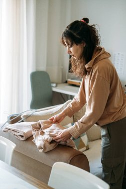 Kadın parlak oturma odasındaki kanepede sweatshirt katlıyor. Sürdürülebilir moda alışkanlıkları, giysi bakımı, kapsül giysi bakımı