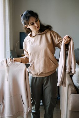 Kadın pencere kenarındaki askıda duran kıyafetlere özenle bakıyor. Önce konfor modası, şezlong giyimi estetik, melez iş stili.