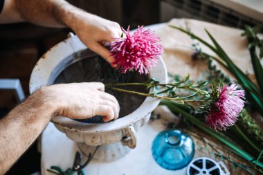 Eller pembe paskalya çiçeğini çalışma masasındaki taş vazonun içine yerleştirir. Geleneksel çiçekçilik teknikleri, el yapımı buketler, özgün yaratıcı süreç.