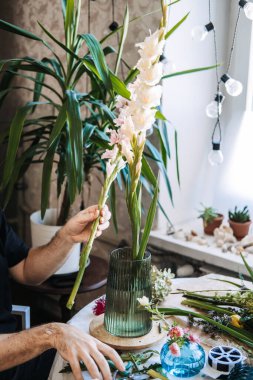 Eller çalışma masasındaki yeşil cam vazonun üzerinde uzun beyaz gladiolus saplarını tutar. Sıfır atık çiçekçi, sürdürülebilir çiçekli alternatifler, çevre dostu çiçek düzenleme
