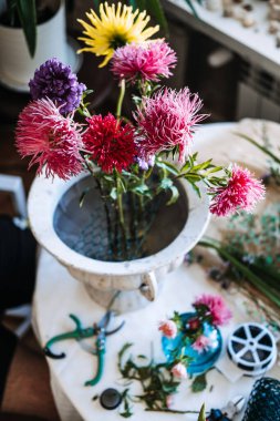 Mor ve pembe paskalya çiçekleri dağınık çalışma masasındaki beyaz taş vazonun içinde tel örgü şeklinde duruyor. Yaratıcı sağlık, bitkiler aracılığıyla stres atma, dikkatli çiçek düzenlemesi.