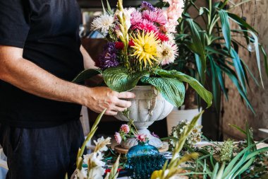 Renkli asterlerin ve büyük yeşil hosta yapraklarının bulunduğu taştan vazodaki çiçek aranjmanlarını tutan eller. Çiçek tasarım sınıfı, DIY çiçek düzenleme, çiçekçilik atölyesi