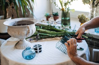 Eller beyaz taş vazonun yanındaki masada yeşil ağları şekillendirmek için tel kesiciler kullanır. Yavaş çiçek hareketi, el yapımı düzenlemeler, çiçek atölyesi.