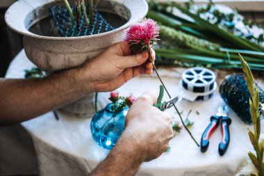Eller, taş vazo ve çiçek tellerinin yanındaki masanın üzerindeki pembe çiçek sapını kesmek için küçük kesikler kullanır. Özenli çiçek düzenleme, botanik meditasyon, duyusal hobiler.